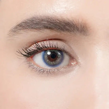 Extreme close-up of a human eye wearing a blue-gray colored contact lens, with long black eyelashes, a defined eyebrow, and shimmering eyeshadow on a pale skin background.